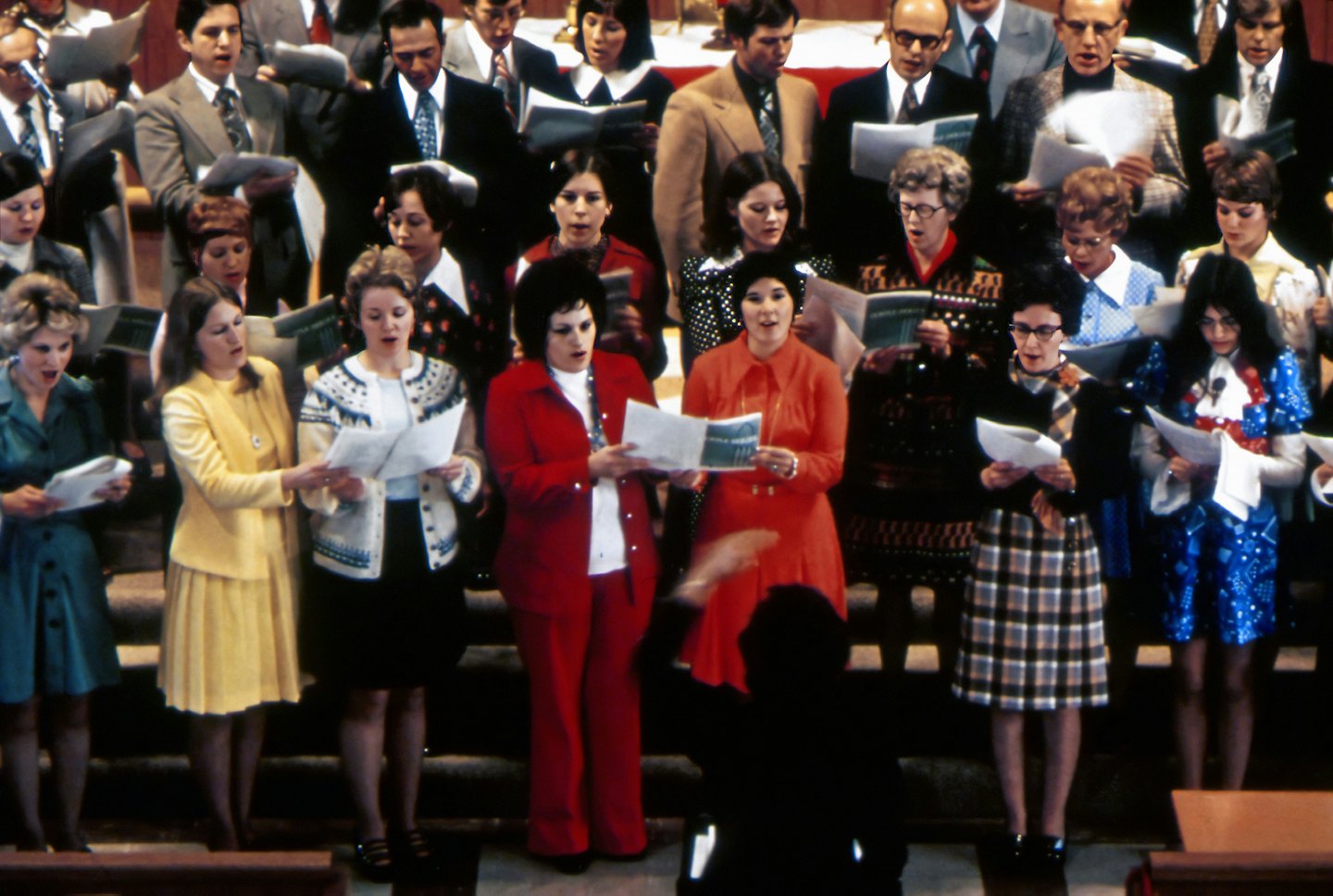 A choir sings from sheet music on stepped platforms directed by a conductor.