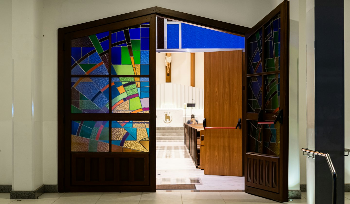 Open dark wooden doors with stained glass leading into a bright church.